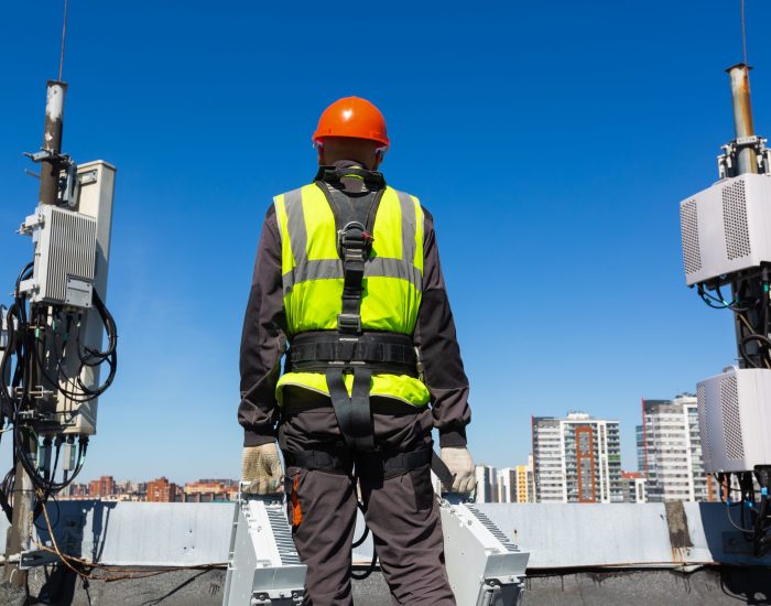Telecommunication engineer  in helmet and uniform holds telecomunication equipment in his hand and  antennas of GSM  DCS UMTS LTE bands, outdoor radio units, optic fibers, power cables are installed on the roof. Working process of upgrading telecommunication equipment